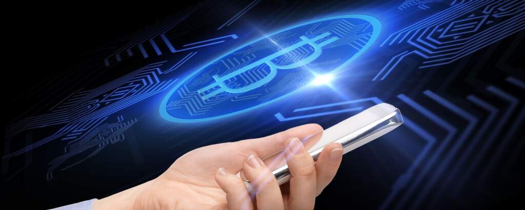 A hand holding a mobile phone in front of a digital hologram of bitcoin, symbolizing the use of blockchain technology and decentralized transactions.